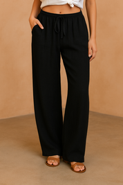 Black linen wide-leg pants with a relaxed drape, elastic drawstring waist, and side pockets, styled with tan flat sandals.