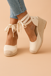 Cream-colored canvas wedge espadrilles with woven jute heels and thick ankle ties, featuring textured toe caps and minimalist stitching.