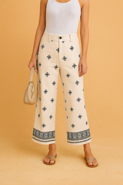 Cream-colored cropped wide-leg pants featuring black geometric embroidery throughout and an ornate black border design along the hem, styled with a button closure, belt loops, and paired with neutral flat sandals.