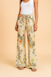 High-waisted wide-leg linen pants in light beige with a vivid tropical floral print in orange, yellow, and olive tones, featuring a drawstring waistband and a relaxed silhouette, paired with neutral flat sandals.