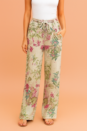 Beige wide-leg linen pants featuring a vibrant botanical floral print in shades of magenta, green, and teal, styled with a drawstring waistband and paired with neutral-tone sandals.