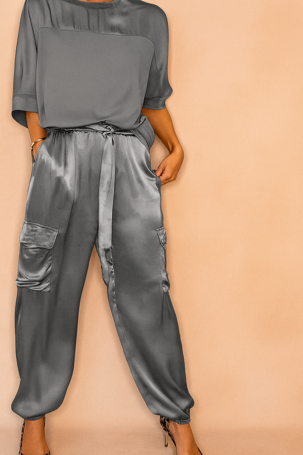 Gray satin cargo pants with subtle camouflage texture, side flap pockets, ankle cuffs, and tie-waist detailing, paired with a matte-finish loose satin blouse and leopard-print heels.