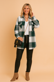 Women's oversized green and grey plaid shacket with dual chest pockets, styled over a white V-neck top and black high-rise leather pants with silver button detailing, completed with tan suede ankle boots and a black crossbody bag.