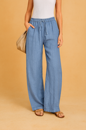 Light blue linen drawstring pants in a relaxed wide-leg silhouette, featuring an elastic waistband and side pockets, styled casually with tan flat sandals and a slouchy beige handbag.