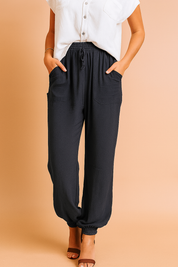 Relaxed-fit navy blue drawstring jogger pants featuring front patch pockets and elastic ankle cuffs, paired with a white short-sleeve button-up blouse and brown open-toe heels.