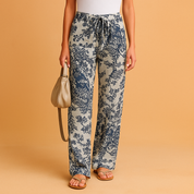 High-rise straight-leg linen pants in a light beige base with bold navy blue floral print, featuring a matching drawstring waistband and relaxed fit, styled with tan flat sandals and a neutral leather handbag.