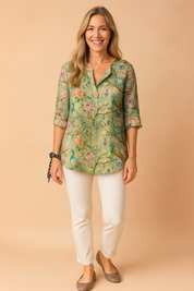 Green vintage-style blouse with vibrant multicolored floral and peacock print, three-quarter sleeves, and a single front button, paired with slim white pants and taupe flats.