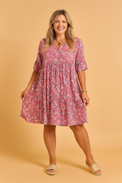 Relaxed-fit pink floral babydoll dress with elbow-length sleeves, gathered waistline, tiered hem, and scattered white and black daisy print.