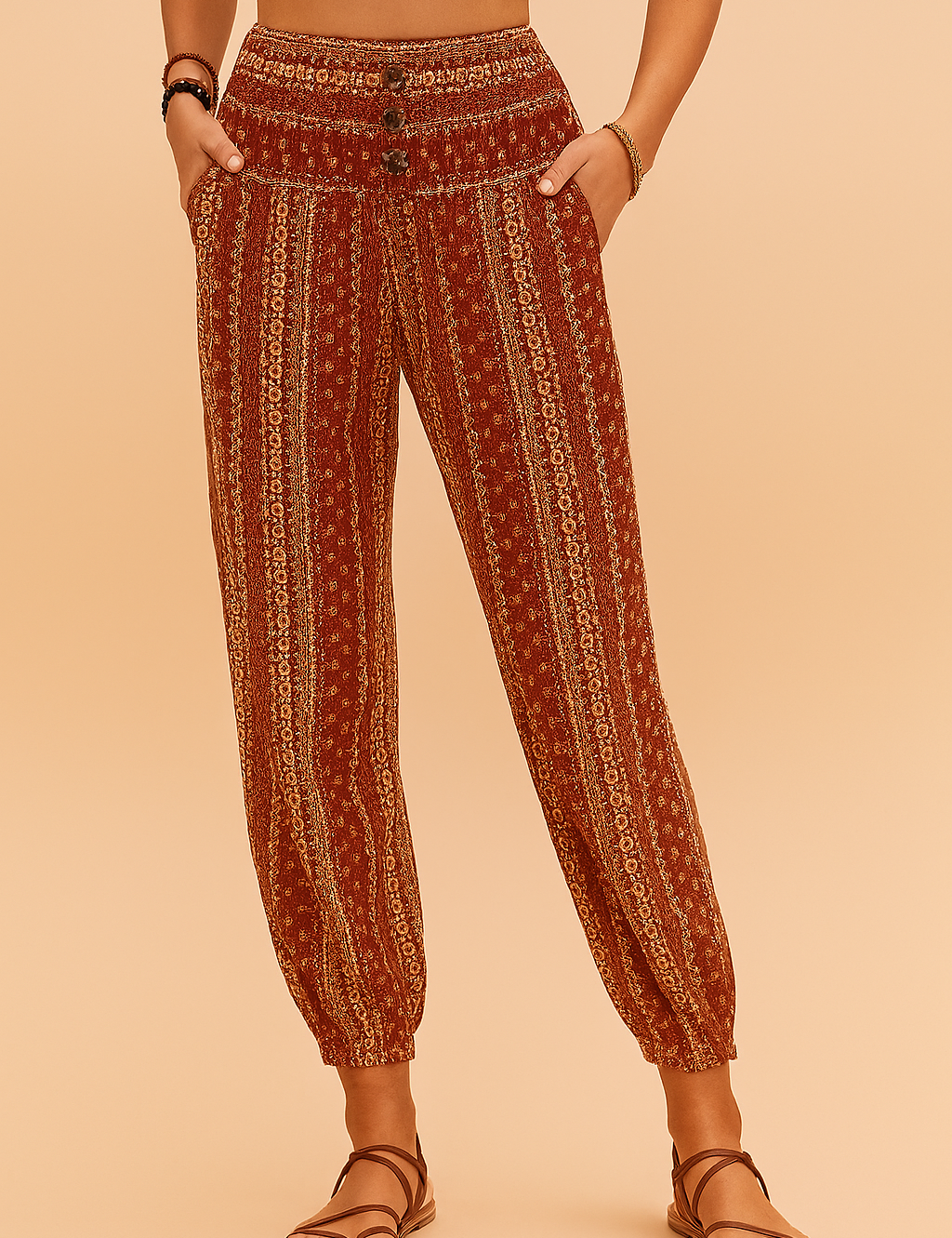 Boho-style high-waisted jogger pants featuring a rust red base with vertical floral and geometric beige prints, smocked waistband with three buttons, side pockets, and elasticated cuffs, paired with brown strappy flat sandals.
