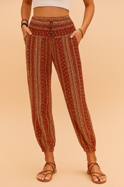 Boho-style high-waisted jogger pants featuring a rust red base with vertical floral and geometric beige prints, smocked waistband with three buttons, side pockets, and elasticated cuffs, paired with brown strappy flat sandals.