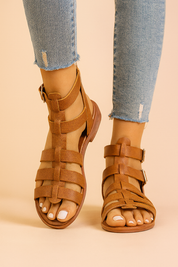 Tan gladiator sandals featuring multiple wide straps, ankle buckle closures, and a flat sole with subtle grain texture.