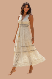 Sukienka boho maxi z delikatnej koronki w odcieniu ecru, z głębokim dekoltem w serek, zdobiona geometrycznymi wzorami i haftowanymi wstawkami w pasie oraz na dole; fason rozkloszowany z ozdobnymi poziomymi przeszyciami i misternym wykończeniem u dołu.