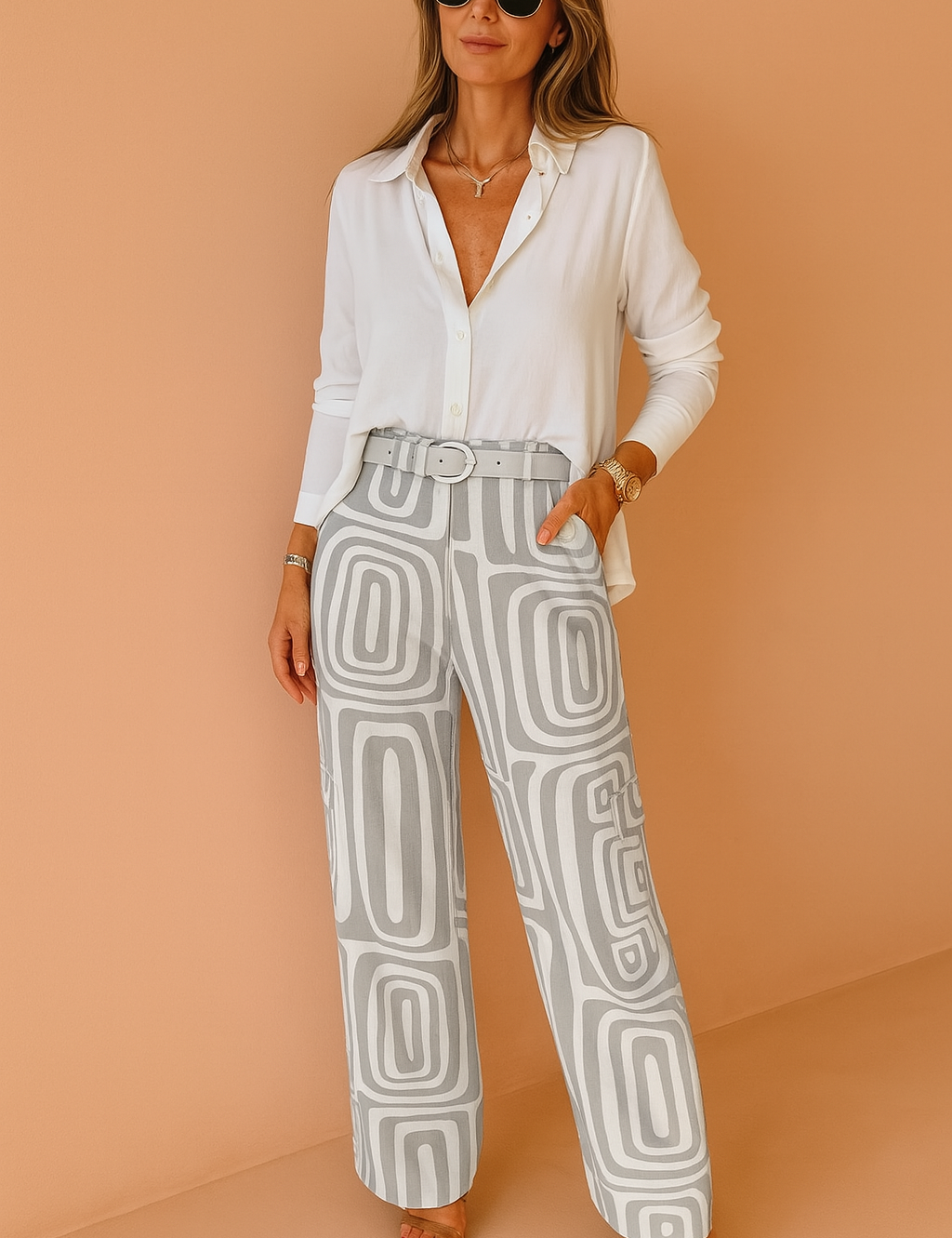 White button-up blouse paired with high-waisted wide-leg gray and white geometric print trousers, styled with a matching belt and brown open-toe heels, against a warm peach studio background.