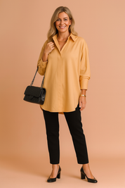 Mustard yellow oversized long-sleeve blouse with a clean collar and concealed front placket, paired with fitted black trousers and black block-heeled pumps; completed with a quilted black chain-strap handbag.