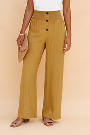 High-waisted mustard wide-leg linen-blend pants with a shirred elastic waistband, front vertical button placket with three dark buttons, and a natural matte texture finish. Styled with a tucked-in white top and taupe block heel sandals.