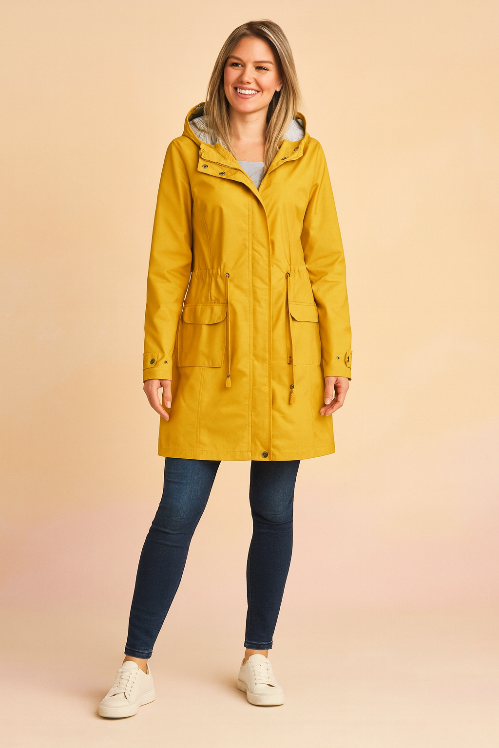 Mustard yellow hooded raincoat with front flap pockets, drawstring waist, and snap-button cuffs, paired with dark blue skinny jeans and white low-top sneakers.
