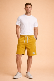 Mustard yellow ribbed corduroy shorts with elastic waistband, white adjustable drawstring, straight knee-length cut, soft vertical cord texture, and woven logo patch on the hem, featuring a relaxed casual fit with identical fabric pattern and color consistency throughout.