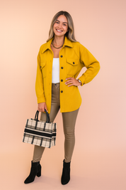 Mustard yellow button-up shirt jacket with front flap pockets, styled with a white tank top, beige fitted leather-look pants, black ankle boots, and a plaid structured handbag.