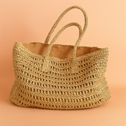 Oversized natural raffia tote bag with open-weave crochet pattern and twin braided handles, styled on a soft sand and pastel pink gradient background.
