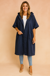 Oversized navy blue hooded coat with short sleeves, single button closure, and two large front pockets, styled with light blue slim-fit jeans and beige suede pumps for a relaxed yet chic look.