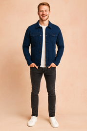 Dark navy blue denim jacket with front button closure, two flap chest pockets, long sleeves, and a straight hem, styled over a plain white t-shirt with slim dark jeans and white sneakers.