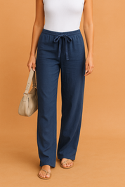 Navy blue wide-leg drawstring pants styled with a white ribbed tank top, beige strappy sandals, and a neutral shoulder bag, set against a warm sandy orange backdrop.