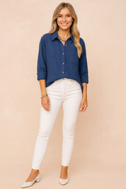 Dark blue button-down linen shirt with a front pocket and rolled-up sleeves, tucked slightly into high-rise white jeans and styled with layered chains and ivory low heels.