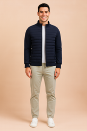 Navy blue quilted zip-up jacket with ribbed cuffs, horizontal padded front detailing, and side pockets, styled over a white shirt and paired with light trousers.