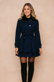 Navy blue double-breasted trench coat with gold button detailing, layered hemline, belted waist tie, and black faux fur collar, styled with sheer black tights and over-the-knee black boots.