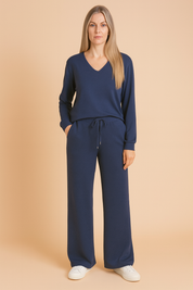 Two-piece navy blue loungewear set featuring a long-sleeve V-neck top and wide-leg drawstring pants made from soft jersey fabric, styled with white sneakers.