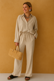 Neutral-toned two-piece co-ord set featuring a long-sleeve button-up shirt and wide-leg drawstring pants, styled with a chunky chain necklace and woven straw handbag for a relaxed yet elegant resort-ready outfit.