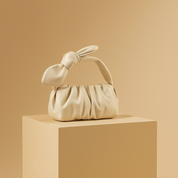 Off-white ruched handbag with a knotted bow handle, placed on a beige pedestal against a soft sand-to-blush gradient background.
