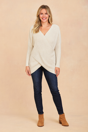 Off-white textured knit crossover sweater with asymmetrical hem, paired with dark-wash skinny jeans and tan suede ankle boots.