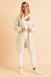 Off-white chunky knit longline cardigan with ribbed texture, relaxed open-front design, and drop-shoulder long sleeves, styled with a white cut-out top, slim white pants, and white sneakers for a cozy yet modern look.