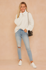 Chunky knit off-white turtleneck sweater paired with light-wash ripped skinny jeans, white sneakers, and a black quilted shoulder bag.