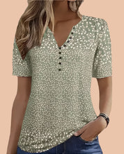 Olive green short-sleeve blouse with a scattered white floral print, V-neckline, and decorative black buttons; casual yet feminine top designed with a soft, lightweight fabric and a flattering hip-length cut.