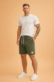 Men’s olive green corduroy shorts with fine vertical ribbed texture, elastic waistband with white drawstring, mid-thigh length relaxed fit, side hand pockets, and stitched rectangular logo patch on the lower hem, featuring a soft matte finish and uniform color tone throughout.