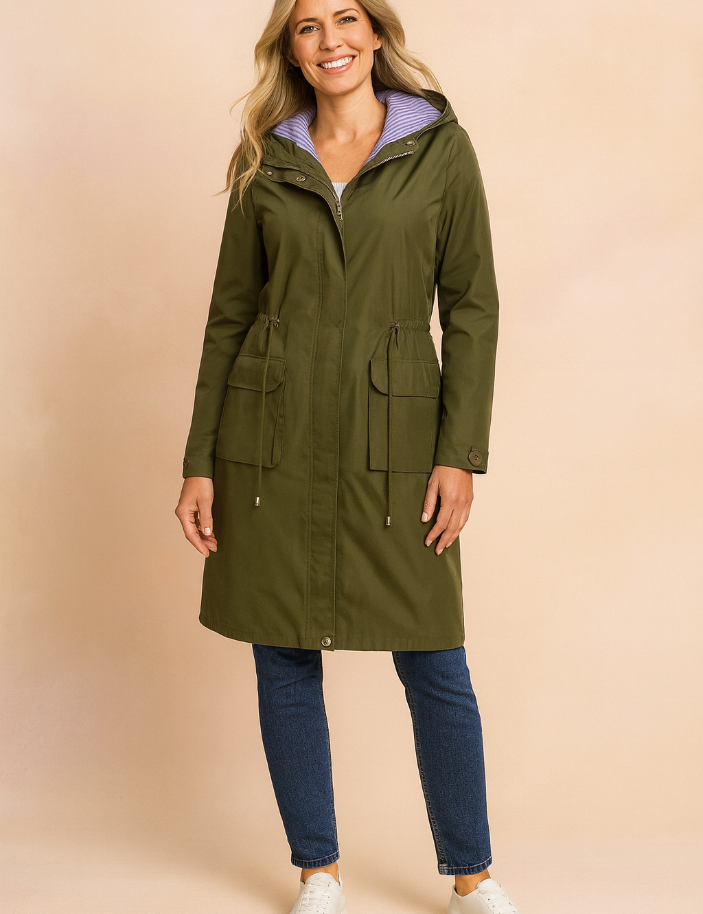 Olive green hooded raincoat with front flap pockets, adjustable drawstring waist, and lavender striped lining, paired with dark blue straight-leg jeans and white low-top sneakers.