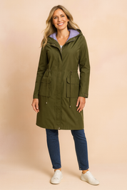 Olive green hooded raincoat with front flap pockets, adjustable drawstring waist, and lavender striped lining, paired with dark blue straight-leg jeans and white low-top sneakers.