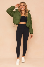 Olive green cropped puffer jacket with front snap buttons, paired with a black sports bra and high-waisted black leggings, styled with white sneakers.