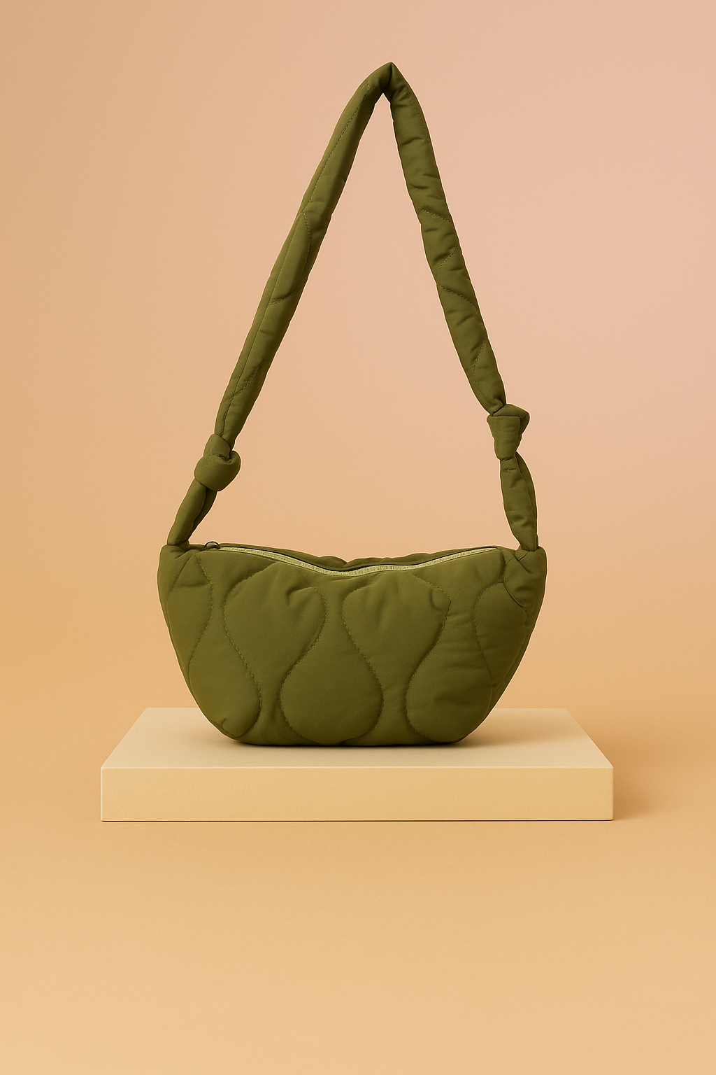 Olive green quilted shoulder bag with crescent shape and knotted padded strap, displayed on a neutral platform with a warm sand and pastel pink gradient background.