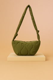 Olive green quilted shoulder bag with crescent shape and knotted padded strap, displayed on a neutral platform with a warm sand and pastel pink gradient background.