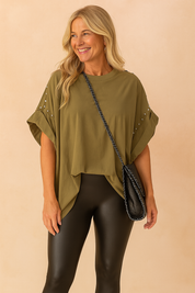Olive green oversized poncho-style top with rolled sleeves featuring silver stud embellishments, paired with black faux leather leggings and a black chain-strap shoulder bag.