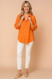  Bright orange long-sleeve button-up crinkled cotton shirt with a relaxed fit, featuring white buttons, paired with fitted white ankle-length trousers and nude pointed-toe heels.