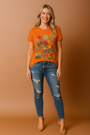 Bright orange T-shirt with intricate multicolor floral print featuring grouped blossoms in various sizes and shades, styled with distressed blue denim jeans for a bold and expressive casual look.