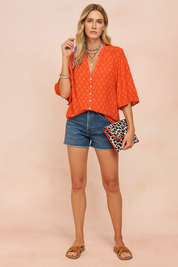 Relaxed-fit orange patterned blouse with white button-down front, styled with casual mid-rise denim shorts and camel cross-strap sandals. The lightweight blouse features three-quarter sleeves and a subtly textured fabric perfect for summer or resort wear, paired with a statement leopard-print clutch.