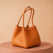 Compact trapezoid-shaped orange leather handbag with dual thin handles and soft pebbled texture, displayed on a muted pink round platform against a warm pastel gradient background.
