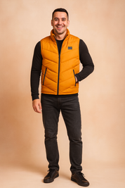 A young man in his mid-to-late twenties stands confidently against a soft beige backdrop, his dark brown hair styled upward at the front and his warm brown eyes framed by subtle laugh lines. He wears a bright orange puffer vest over a black fitted shirt and dark gray jeans, complete with low-top sneakers, while striking a relaxed pose with a casual smile and a confident, direct
