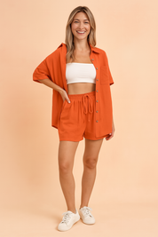 Vibrant orange two-piece coord set featuring a relaxed-fit button-up shirt with short sleeves, a chest pocket, and matching high-waisted drawstring shorts with visible patch pockets. The fabric has a smooth, solid weave and consistent color throughout, maintaining the original shade and texture precisely.