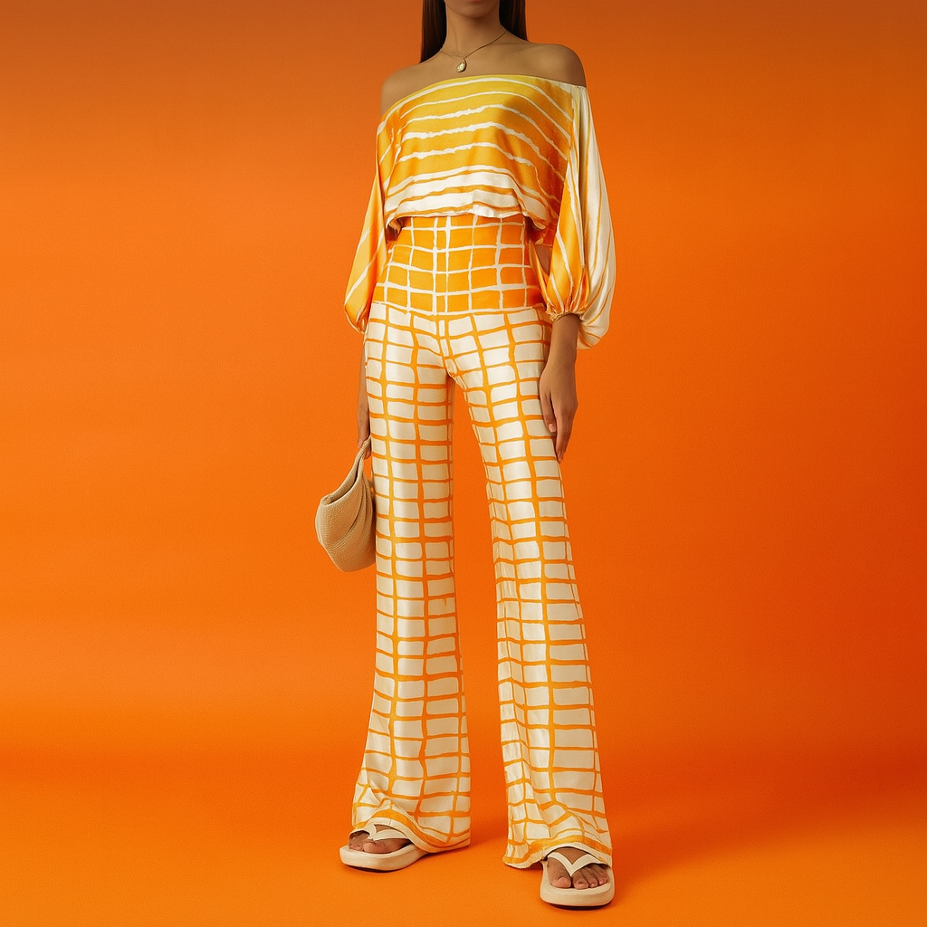Golden-orange tie-dye coord set featuring a wide boat-neck blouse with voluminous sleeves and high-waisted wide-leg pants with grid print, styled with platform sandals and a beige woven handbag on a vibrant orange studio background.